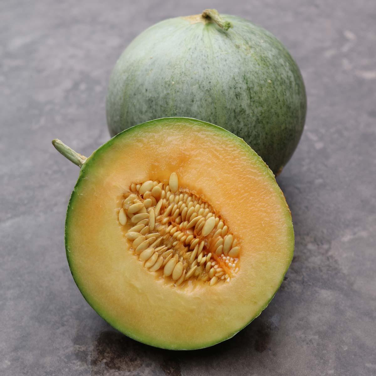 Whole and halved Charentais melon on a stone surface, showing its smooth gray-green rind and vibrant orange flesh with seeds